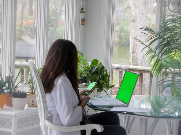 A person is sitting at a desk working on a laptop and filling out a chart on a notebook. There are charts and graphs taped to the wall. The room is bright and professional and has lots of plants.